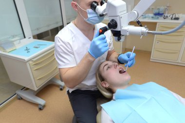 Erkek bir dişçi, bir hastanın dişlerini profesyonel bir diş mikroskobuyla inceler. Müşteri dişçi koltuğunda yatıyor..