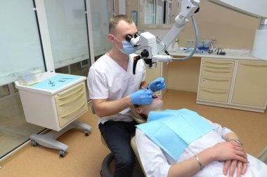 Erkek bir dişçi, bir hastanın dişlerini profesyonel bir diş mikroskobuyla inceler. Diş hekimi yenileme aletleri ile taşıyıcıları engeller. Müşteri dişçi koltuğunda yatıyor..