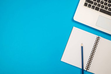 Not defterinde kalem, izole edilmiş mavi arkaplanda dizüstü bilgisayarı olan iş yerinin üst görünüm fotoğrafı..