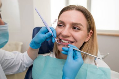 Maskeli bir dişçi ve bir hasta, gülümseyen genç sarışın bir hastayı tedavi eden bir dişçi. Diş hekimliği, modern diş sağlığı kliniği kavramı.