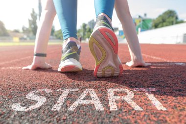 A woman is waiting for the start on an athletics track with the word start engraved on the floor. Large plan of the legs of an athlete at the start