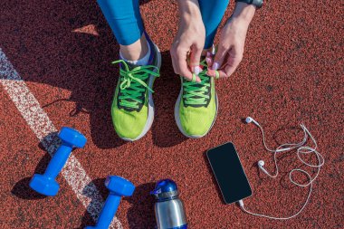 female runner tying her shoe laces, getting ready to race on a stadium running track with a bottle of water and a phone in headphones.View from above.