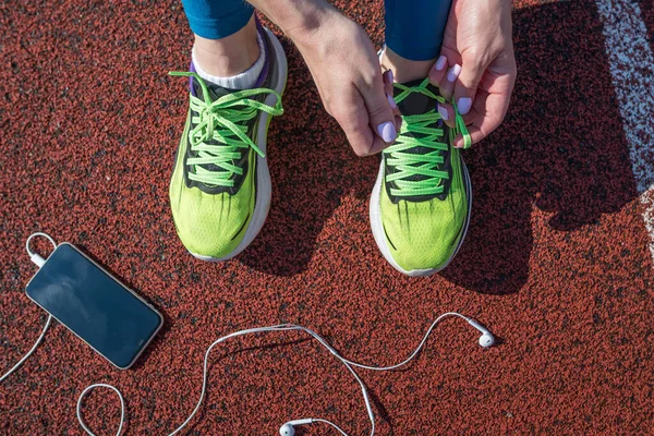 female runner tying her shoe laces, preparing for a race on a stadium running track with a phone in her headphones
