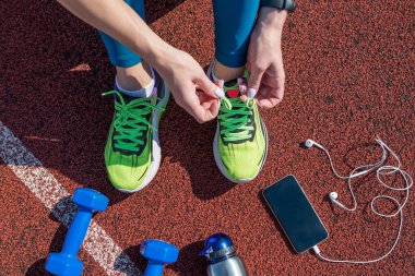female runner tying her shoe laces, getting ready to race on a stadium running track with a bottle of water and a phone in headphones.View from above.
