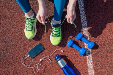 female runner tying her shoe laces, getting ready to race on a stadium running track with a bottle of water and a phone in headphones.View from above.