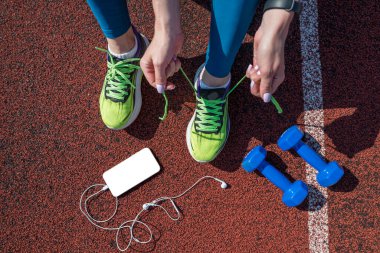 female runner tying her shoe laces, preparing for a race on a stadium running track with a phone in her headphones