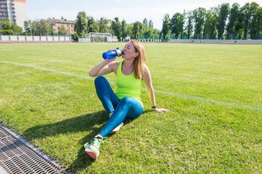 Enerji, canlılık, refah konsepti. Futbol sahasında oturan formda sporcu kadın, elinde sallayıcı, içme suyu ya da protein, açık hava egzersizlerinden sonra mola veriyor..
