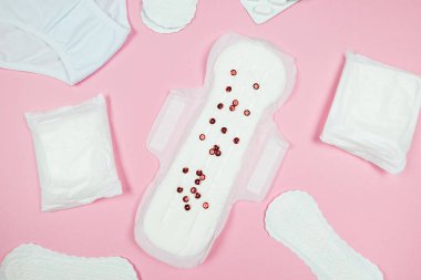 Top view photo of sanitary napkins with red confetti, underwear, pain pills on isolated pastel pink background
