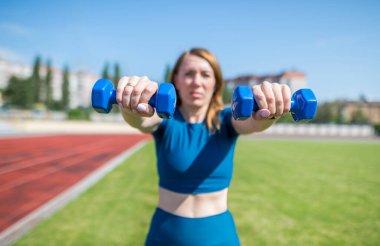 Sport and fitness concept. A healthy lifestyle. Young female athlete with dumbbells in her hands at the stadium. A woman does exercises with dumbbells at a sports stadium.