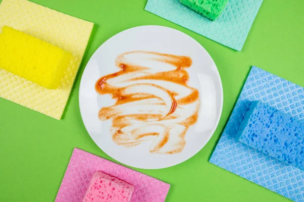 Photo from above of a dirty plate and multicolored cleaning pads on an isolated green background