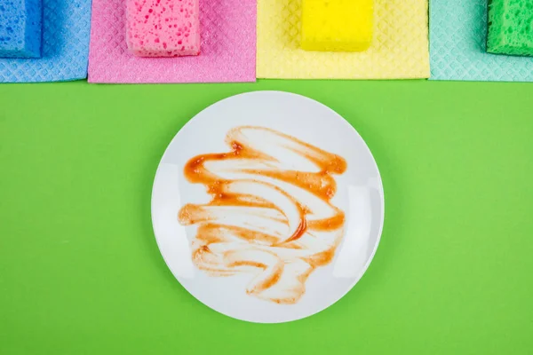 Photo from above of a dirty plate and multicolored cleaning pads on an isolated green background