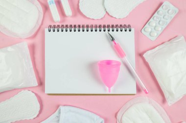 Top view photo of open planner over sanitary napkins, tampons, underwear and pills on isolated pastel pink background with blank space