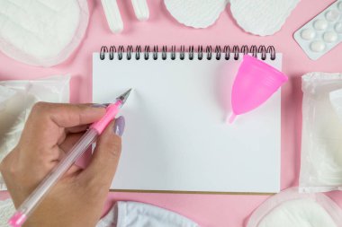 Pembe arka planda beyaz boş bir not defterine yazan bir kadının el resmi. Kadınların adet görme konsepti, kistik günler..