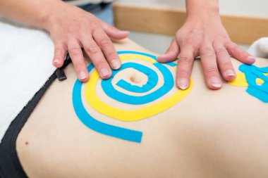 Kadın bir fizyoterapistin elleri, bir doktorun ofisindeki kanepede yatan genç, ince, atletik bir kadının karın kaslarına yapışkan elastik tedavi bandı uyguluyor. Yaralı Rehabilitasyon ve Sağlık Hizmetleri