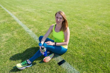 Spor giyimli atletik bir kadın stadyumdaki çimlerin üzerinde oturuyor.