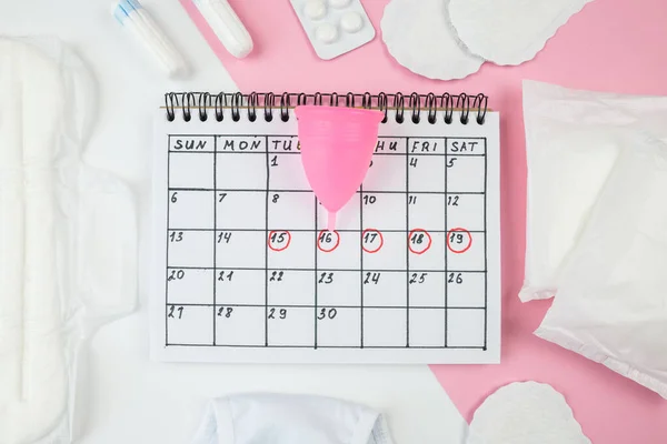 Top view photo of red calendar marks, panties, menstrual cup, sanitary pads and tampons on isolated white and pastel pink background
