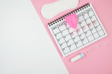Beyaz ve pembe arka planda, kadınların kritik günlerinin pedleri ve aksesuarları olan takvimin üst görüntüsü. Kadın adet dönemi kavramı..