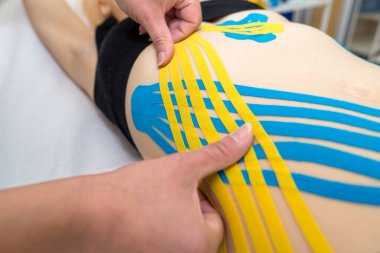 Kadın bir fizyoterapistin elleri, bir doktorun ofisindeki kanepede yatan genç, ince, atletik bir kadının karın kaslarına yapışkan elastik tedavi bandı uyguluyor. Yaralı Rehabilitasyon ve Sağlık Hizmetleri