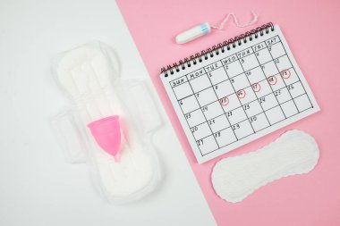 Beyaz ve pembe arka planda, kadınların kritik günlerinin pedleri ve aksesuarları olan takvimin üst görüntüsü. Kadın adet dönemi kavramı..