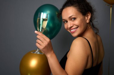 Çekici zarif bir kadın gri duvarın karşısında duruyor ve gülümsüyor, omzunun üzerinden kameraya bakıyor, altın ve metalik yeşil hava balonlarına karşı şampanya flütüyle poz veriyor.