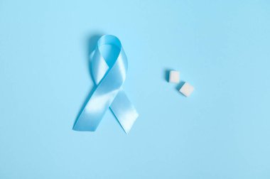 Dünya Diyabet Farkındalığı Günü 14 Kasım 'ın sembolü, saf rafine beyaz şeker küpleri ve mavi saten kurdelelerden oluşan düz bir kompozisyon. Renkli mavi arkaplan üzerinde izole edilmiş, tıbbi reklam için kopyalama alanı