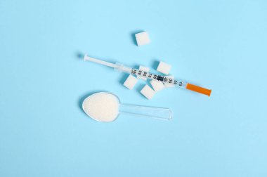 Şeffaf ölçüm kaşığında dağınık beyaz şekerin üst görüntüsü ve mavi zemin üzerinde saf saf şeker küpleri üzerinde insülin şırıngası. Aşırı beyaz şeker yemek hastalık riskini artırır