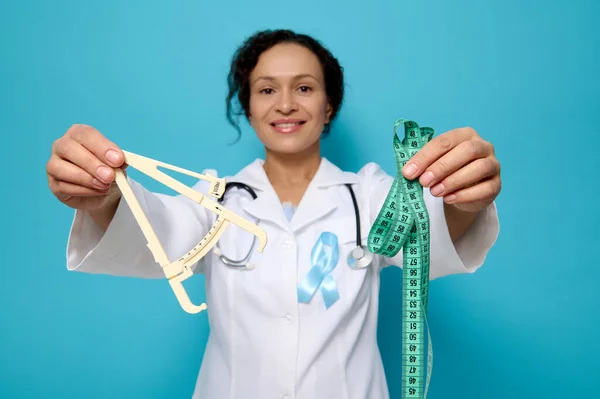 Health care and medical concept, metabolic syndrome disease. Soft focus on caliper and measuring tape in the hands of female doctor nutritionist endocrinologist wearing blue diabetes awareness ribbon.