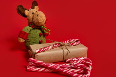 Noel eşyalarının minimalist güzel kompozisyonu, kraft ambalaj kağıdına sarılı hediye kutusu, tatlı lolipoplar ve kırmızı arka planda reklam için fotokopi alanı olan geyik pelüş oyuncaklar.