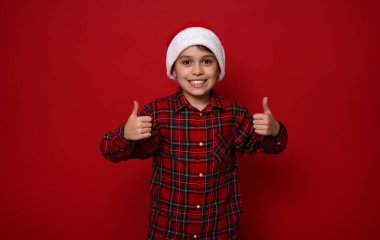 Kırmızı kareli tişörtlü, dişlek gülüşlü Noel Baba şapkası takan ve renkli arka planda fotokopi çekilen kameraya bakan sevimli ergen çocuk.