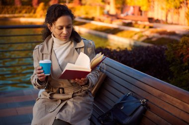 Sıcak, rahat kıyafetli genç bir kadın kitap okuyor ve kahve içiyor. Sonbahar parkındaki ahşap bir bankta oturuyor. Gölün arka planında, suya dökülen sarı yapraklar yansıyor.