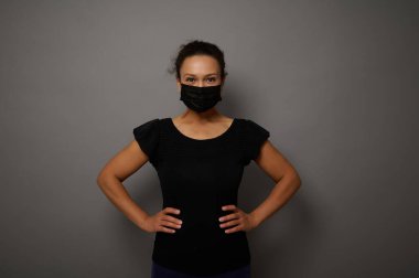 Siyah tıbbi maske takmış, elleri belinde kameraya bakan bir kadının gri arka planda izole edilmiş portresi. Reklam için boşluk kopyala. Kara Cuma konsepti