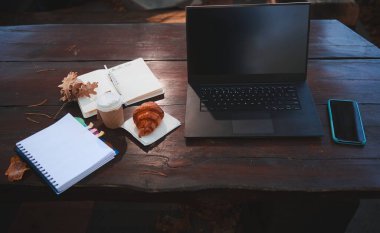 İş ve ofis kırtasiyesinin bileşimi. Laptop, akıllı telefon, açık defter, not defteri ve taze pişmiş kruvasan. Tahta masa yüzeyinde, üzerine düşmüş meşe yaprakları olan bir bardak sıcak içecek.