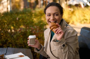Çekici, neşeli bir kadın güneşli bir sonbahar gününde kırsal kesimde kahvaltı ederken kameraya bakıp gülümsüyor. Güzel bayan pişmiş kruvasan yer ve paket kağıttan kahve içer.