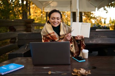 Güzel bir kadın boş bir sayfada kalemle dizüstü bilgisayardaki video konferans sırasında yapılacaklar listesiyle kendini yün ekose battaniyeye sarıp dışarıda ahşap bankta oturarak ısınıyor.