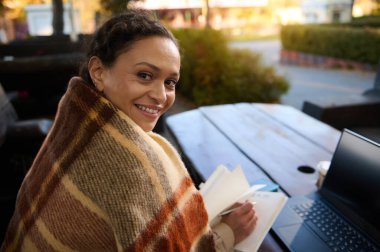 Not defterine yazan neşeli Afrikalı kadın dizüstü bilgisayarla kameraya bakıp gülümsüyor. Karantina süresince e-öğrenme, uzaktan çalışma ve uzak iş kavramı.