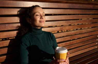 Tahta bir bankta oturan, sırtüstü uzanmış, sevimli, barışçıl genç bir kadının yakın çekimi. Elinde sıcak bir içecek, aletlerden ve şehir telaşından dinlenen, güneşli bir günün keyfini çıkaran bir kağıt bardak tutuyor.