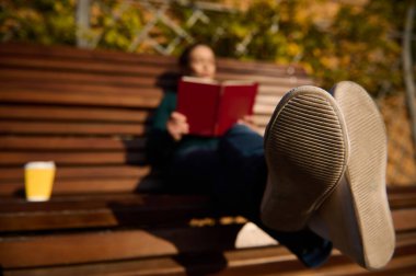 Parktaki ahşap bir bankta dinlenen, kitap okuyan ve kahve içen bulanık bir kadının spor ayakkabılarına odaklanın. Sonbahar doğa arka planında hafta sonunun tadını çıkarıyorum.