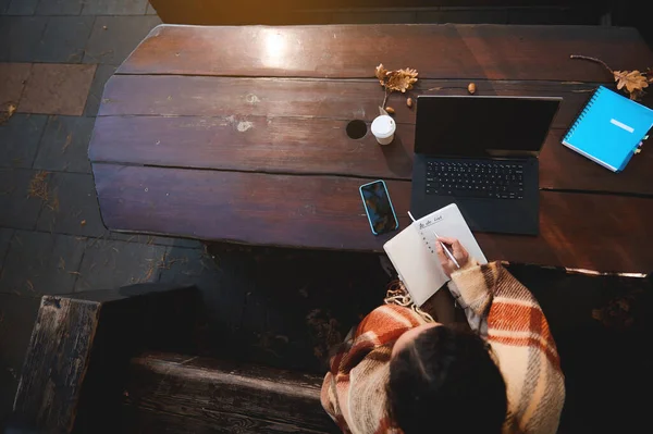 ノートパソコンやオフィス用品の前の木製のベンチに座っている間にノートに書いている女性のオーバーヘッドビュー 隔離閉鎖中のEラーニング リモートワーク リモートビジネスの概念 — ストック写真