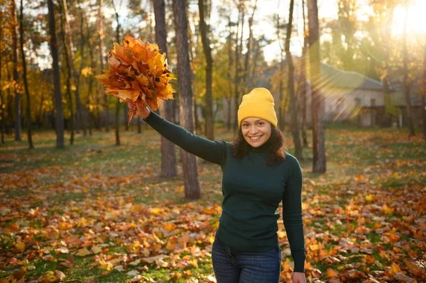 Cheerful mature woman raising hands with collected dry bouquet of fallen autumnal maple leaves and cutely smiling with beautiful toothy smile posing against autumn park with falling sun rays at sunset