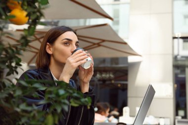 Kısa saçlı, esmer, çekici bir kadın paket kâğıt bardaktan sıcak bir içecek içiyor. Önünde mandalina süsleme ağacı olan rahat bir kafede oturuyor ve kahve molasının tadını çıkarıyor.