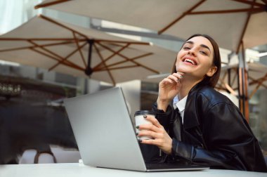 Çekici, modaya uygun, serbest yazar, şık saç kesimi olan iş kadını, yazlık terasta dinleniyor, kahve içiyor, dizüstü bilgisayarda çalışıyor, kameraya güvenle gülümsüyor ve gülüyor.