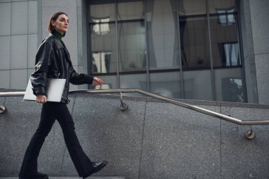 Modern, başarılı, motivasyonlu, kendine güveni tam genç bir kadının portresi elinde dizüstü bilgisayarla modern şehrin caddesinde yürüyor. İş, finans, iletişim, uzaktan çalışma kavramı