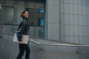 Başarılı motivasyonlu, kendine güvenen kadın serbest çalışan elinde dizüstü bilgisayarla modern yüksek katlı iş merkezinin merdivenlerinden iniyor. İş, finans, iletişim, uzaktan çalışma kavramı