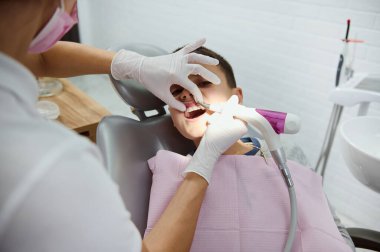 Modern beyaz diş kliniğine pediatrik diş muayenesi sırasında dişçi koltuğunda oturan yakışıklı ilkokul çocuğu tıbbi ağız boşluğu tedavisi görüyor. Diş sağlığı konsepti