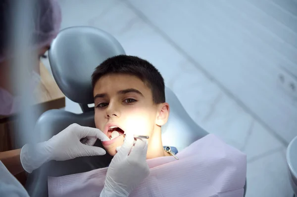 歯科医の椅子に座っている10代の少年の口腔を調べる小児歯科医のオーバーヘッドビューは 歯科ミラーを使用してケアの存在の歯を調べます 歯医者の学校の少年 — ストック写真