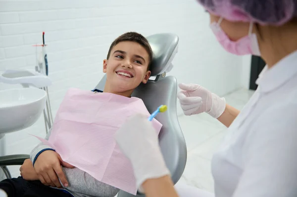 歯科医の椅子に座ってハンサムな男の子は笑顔で美しいおもちゃの笑顔で彼を教えている小児歯科医を見て彼について口腔衛生白クリーン現代医療キャビネットで任命中に — ストック写真
