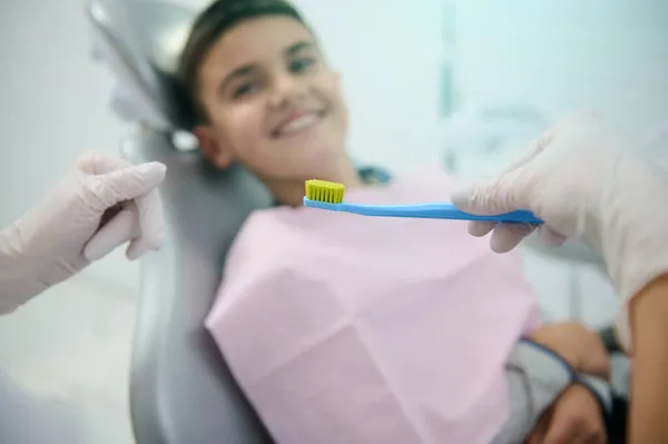 专注于医生手中的牙刷教一个不专注的微笑着坐在牙医椅子上的英俊男孩关于口腔护理和牙齿的日常卫生 医疗概念 — 图库照片