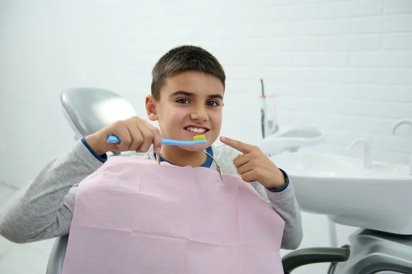 可愛い未成年の少年は 歯科医の椅子に座って 彼の口の近くに歯ブラシを保持するカメラを見て 健康な白い歯で彼の美しい歯の笑顔に指でポイントします 口腔衛生の概念 — ストック写真