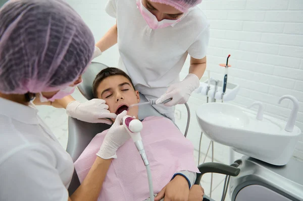 School boy sitting on dentist's chair receiving medical treatment of his oral cavity by pediatrician dentist and his assistant in modern dentistry clinic