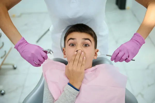 Top view of afraid child boy covering his mouth from fear with hand, sitting on dentist's chair on the background of hands in pink surgical gloves holding stainless steel dental tools near his face